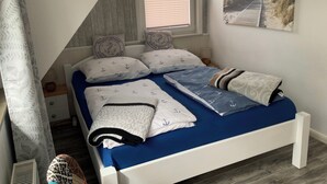 Une chambre avec un plafond en pente, une fenêtre avec des stores, un lit avec une literie bleue et des coussins à motifs d’ancre, et un tableau sur le mur dont le thème est la marine.