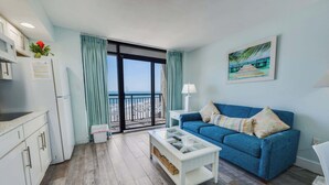 Ein modernes Wohnzimmer mit einem blauen Sofa, einem Glastisch, einer Küchenzeile und einem Balkon mit Blick.