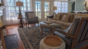 A spacious living room with a large sofa, a chair, and a coffee table. There are two windows with curtains, a fireplace, and a rug on the floor.