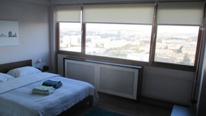 Un dormitorio con cama, radiador y vistas a la ciudad a través de amplios ventanales.