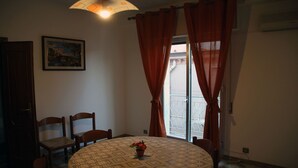 A dining area with a round table, chairs, a vase with flowers, a painting on the wall, and a window with curtains. - Vacation House "La Tana" (Isnello)