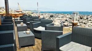 Outdoor wicker furniture arranged on a wooden deck overlooking a rocky shoreline with a view of the sea and distant mountains. - SICILY ETNA SEA Beach&Pool Catania Taormina Etna District (Mascali)