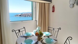 Una mesa de comedor con platos azules y cubiertos, un florero con flores, y vista a un pueblo costero a través de una ventana.