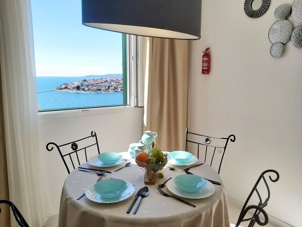 Une table à manger avec de l’argenterie et de la vaisselle bleue, un vase rempli de fleurs et, par la fenêtre, une vue sur une ville côtière.