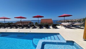 Un área de piscina con sillones de descanso y sombrillas, con vista despejada al mar y piscina con baldosas.