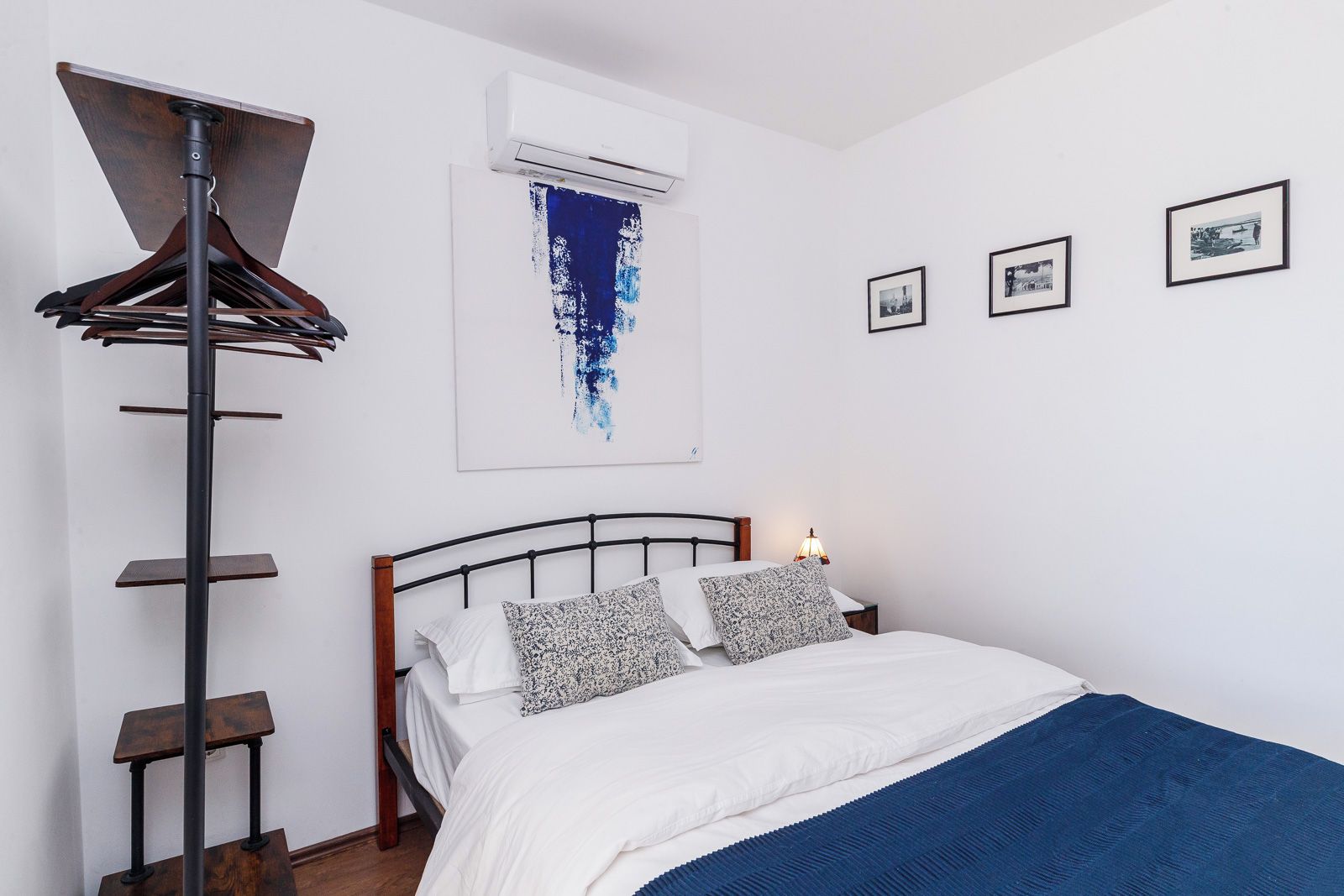Un dormitorio con cama, aire acondicionado de pared, perchero de madera y tres cuadros en la pared.