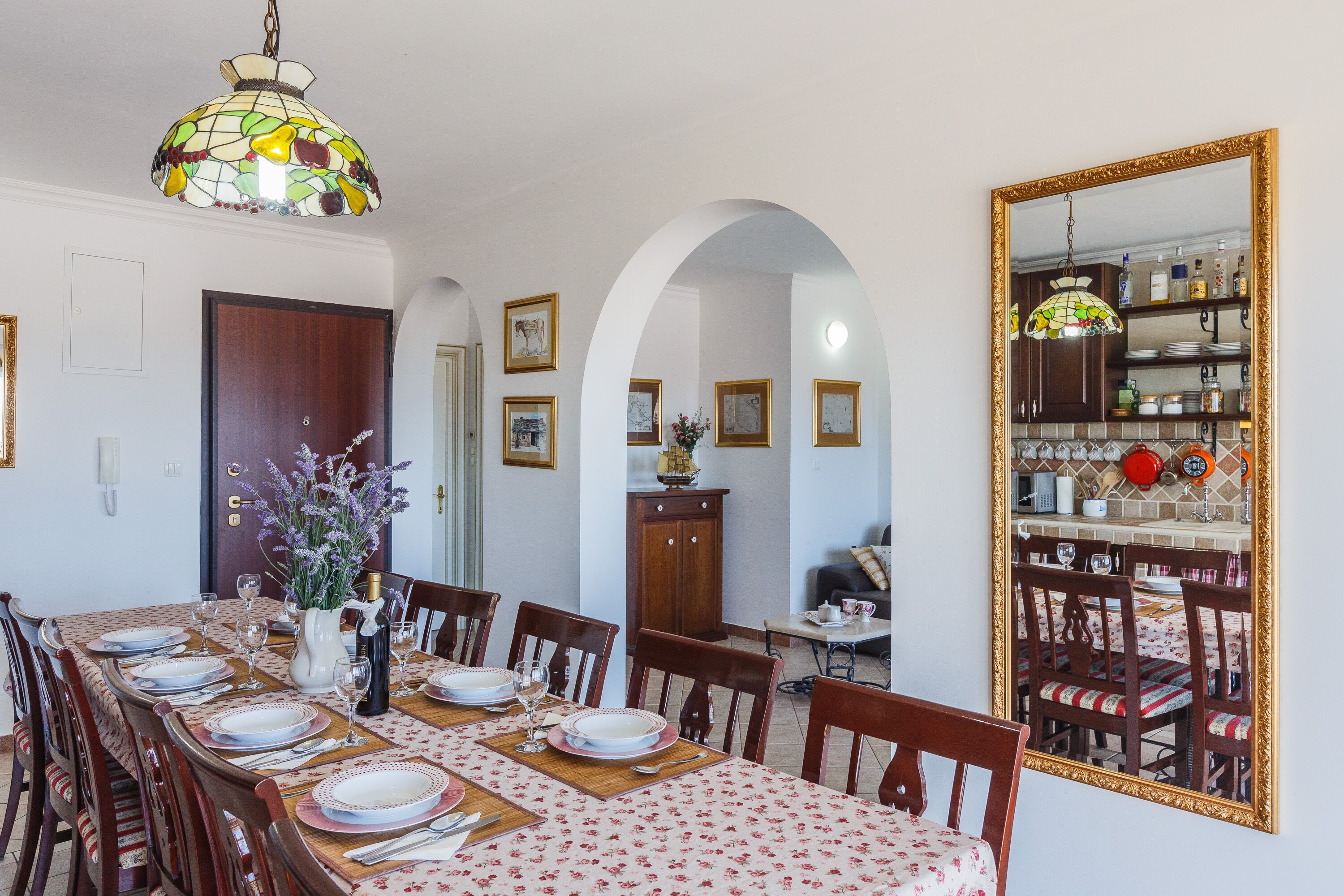 Comedor con una mesa grande dispuesta para seis personas, una araña y un espejo que refleja la cocina.