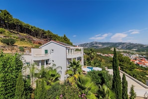 A two-story building with a balcony, surrounded by lush greenery and a mountainous backdrop.