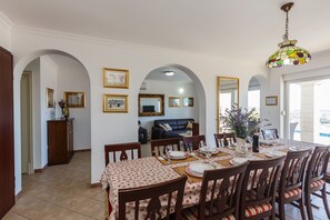 A dining area with a table set for four, surrounded by wooden chairs with cushioned seats. There are framed pictures on the walls, a dark-colored sofa in the background, and a chandelier above the table. -  Idyllic Villa near Split, pool, garden, free parking and stunning view, (OWNER) (Solin)