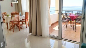 A dining area with wooden chairs and a table, a sliding glass door leading to a balcony with a red tablecloth, and a view of the sea.