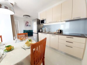 A modern kitchen with a dining area. It includes a refrigerator, microwave, toaster, and a sink. There is a dining table with chairs and a sofa in the background.