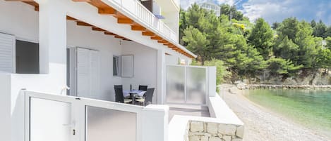 Een modern gebouw met twee verdiepingen, een balkon, een stenen muur en uitzicht op een strand en bomen.