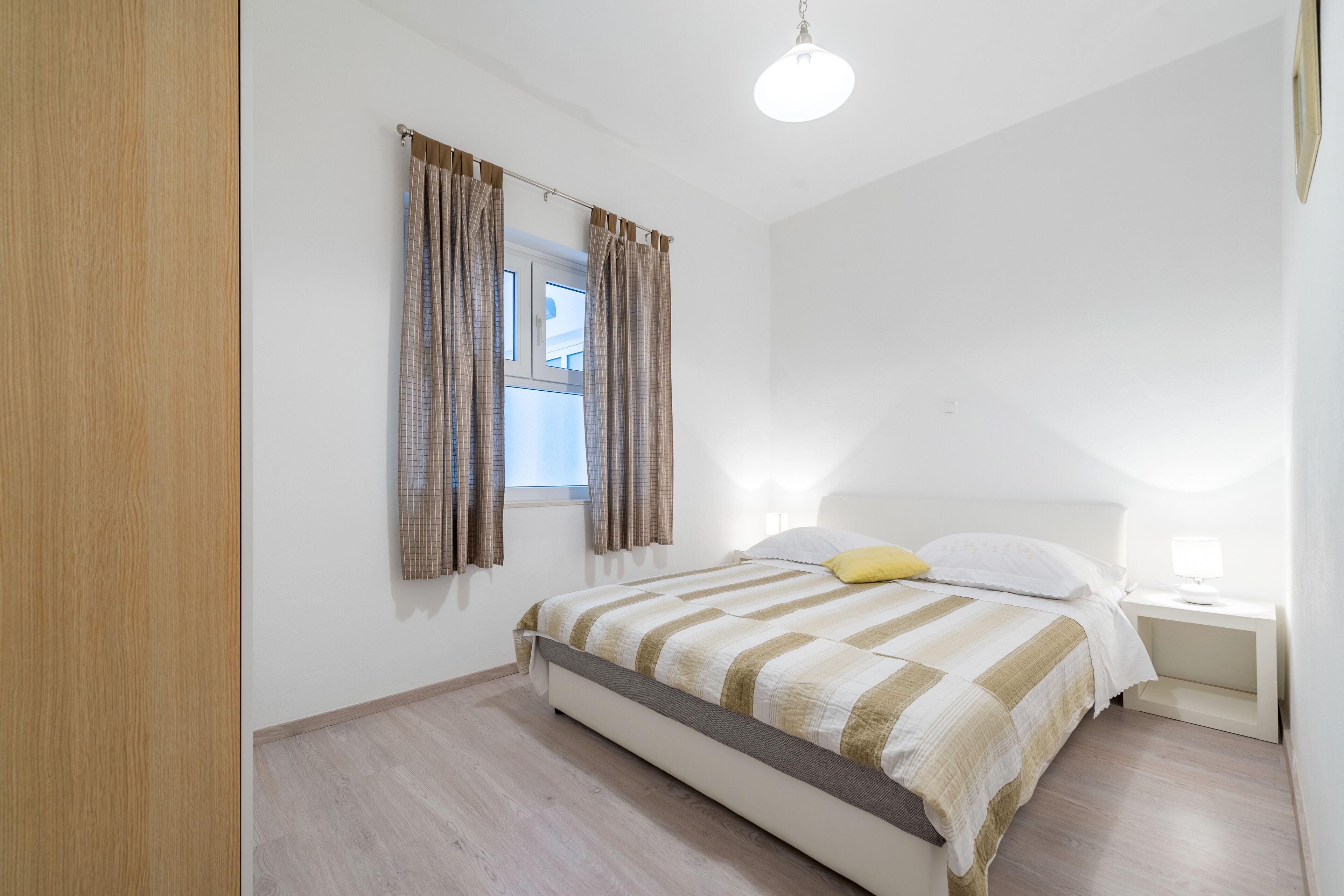 Ein Schlafzimmer mit Bett, Vorhängen, einem Fenster, einem Nachttisch mit Lampe und Deckenleuchte.
