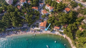 An aerial view of a coastal town with a beach, clear turquoise waters, and a mix of residential buildings with red-tiled roofs.