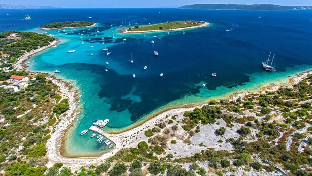 Vista aérea de uma área costeira com águas claras, barcos e uma pequena ilha com edifícios.