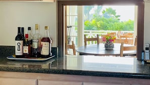 Una cocina con una encimera que muestra varias botellas de licores, una zona de comedor con mesa y sillas, y vistas al exterior con vegetación.