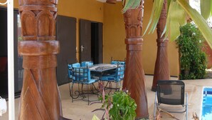 A covered outdoor seating area with a table and chairs, surrounded by large wooden pillars and tropical plants. - Charming recent air-conditioned 3 bedroom villa with swimming pool  (Ngaparou)