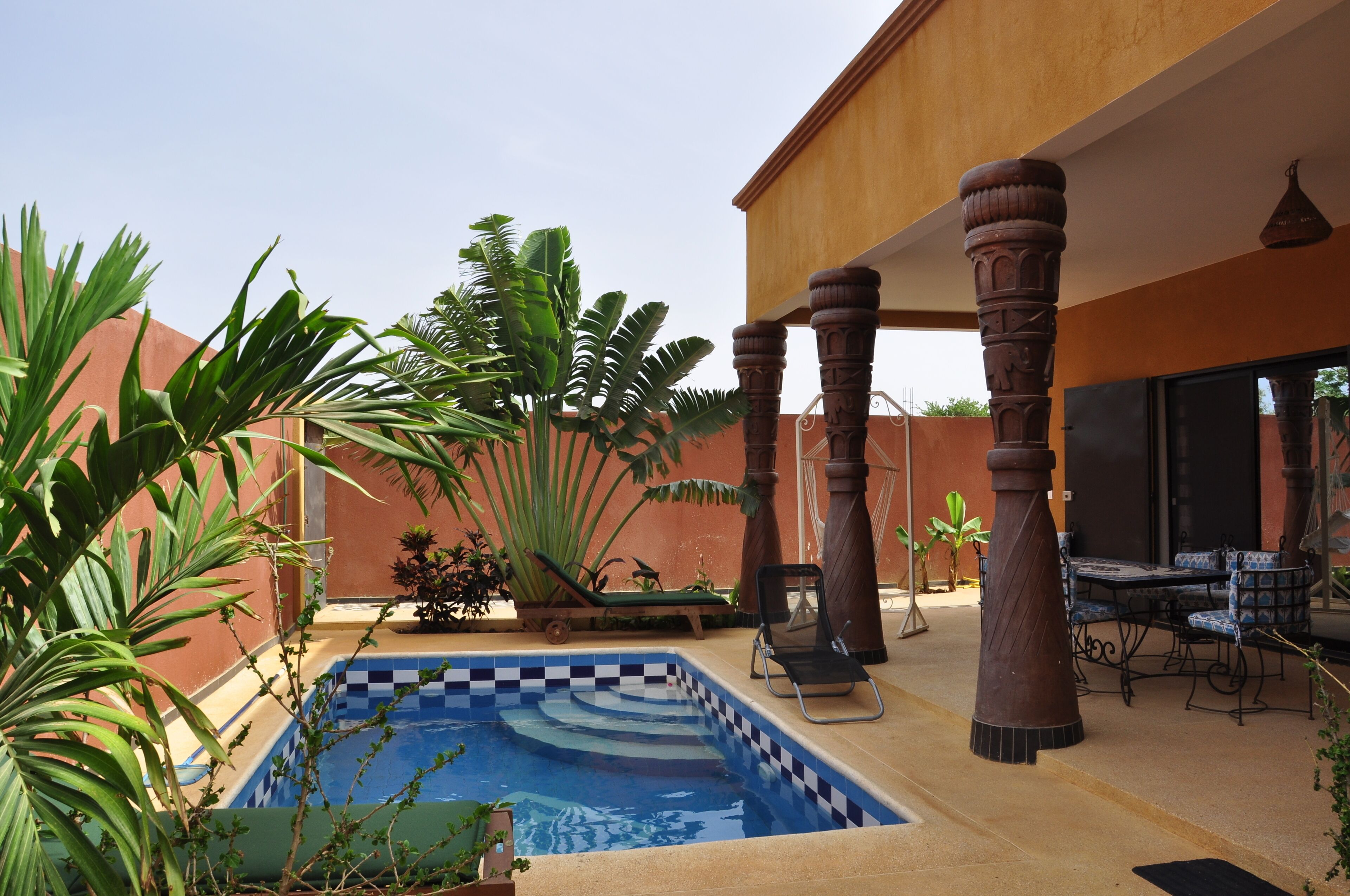 A pool area with a checkered tile edge, surrounded by plants and a wooden pergola.