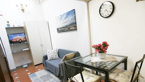 A compact living space with a sofa, a glass-top table with chairs, a clock, a television, and a cityscape painting. - Santa Croce Studio in the historic center (Firenze)