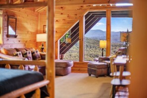 A cozy wooden cabin interior with a large window offering a view of a forested mountain range.