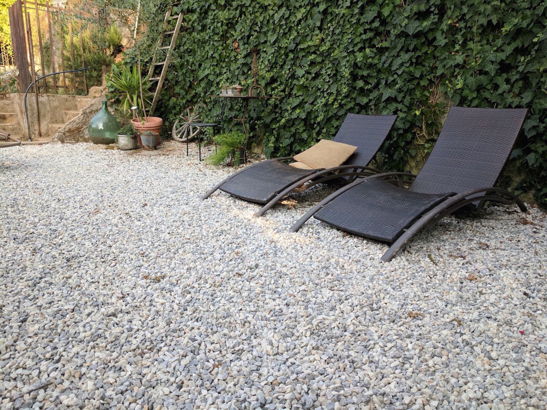Two outdoor lounge chairs with a gravel ground, a wooden ladder, and a wall covered in ivy.