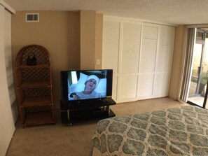 Habitación de hotel con televisor, cubrecama estampado, silla de mimbre y puerta corrediza de vidrio.