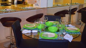 Een eethoek met een glazen tafel gedekt voor vier personen, groene borden en een 'WELCOME' bord boven de balie.