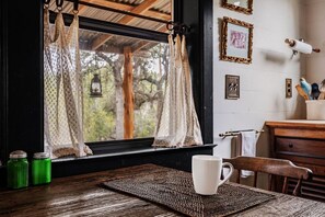 Une table en bois rustique, sur laquelle repose une tasse blanche, avec des rideaux en dentelle et une vue sur les arbres par la fenêtre.