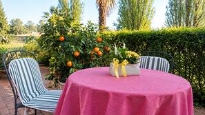 Un área de comedor al aire libre con una mesa redonda cubierta con un mantel rosa, dos sillas a rayas y una maceta con flores amarillas.