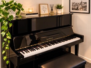 Un piano vertical Kawai negro con un banco, rodeado de objetos decorativos como libros, una vela y una cita enmarcada.