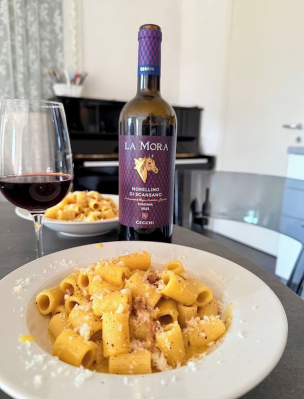 Una botella de vino La Mora Morellino di Scansano y un plato de pasta con una copa de vino tinto.