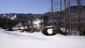A large, two-story house with a balcony, surrounded by snow-covered ground and trees, with mountains in the background.