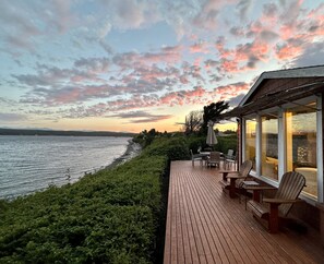 Une terrasse en bois avec des sièges extérieurs, donnant sur une étendue d’eau et un ciel au coucher du soleil.