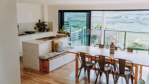 A modern kitchen with a large island, a dining area with wooden chairs, and a large window offering a view of the countryside. - 5-Bed Coastal Retreat w/ Studio, Sauna & Sea Views (Newgale)