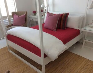 Un letto con struttura bianca e biancheria rossa, un comodino con una lampada e una sedia con un cuscino rosa.
