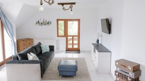 Ein modernes Wohnzimmer mit einem dunkelgrauen Sofa, einem blauen Hocker, einer Holzbalkendecke mit Seillampen und einer Holzkassette sowie einem an der Wand montierten Fernseher.