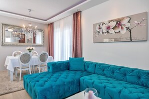 A living room with a teal sectional sofa, a dining area with a white table and chairs, a large mirror, and a floral painting on the wall.