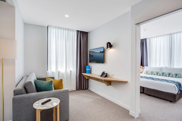 Una habitación de hotel moderna con un sofá gris, una mesita redonda con un control remoto, una cama con colcha estampada, un televisor montado en la pared y un teléfono también sujeto a la pared.
