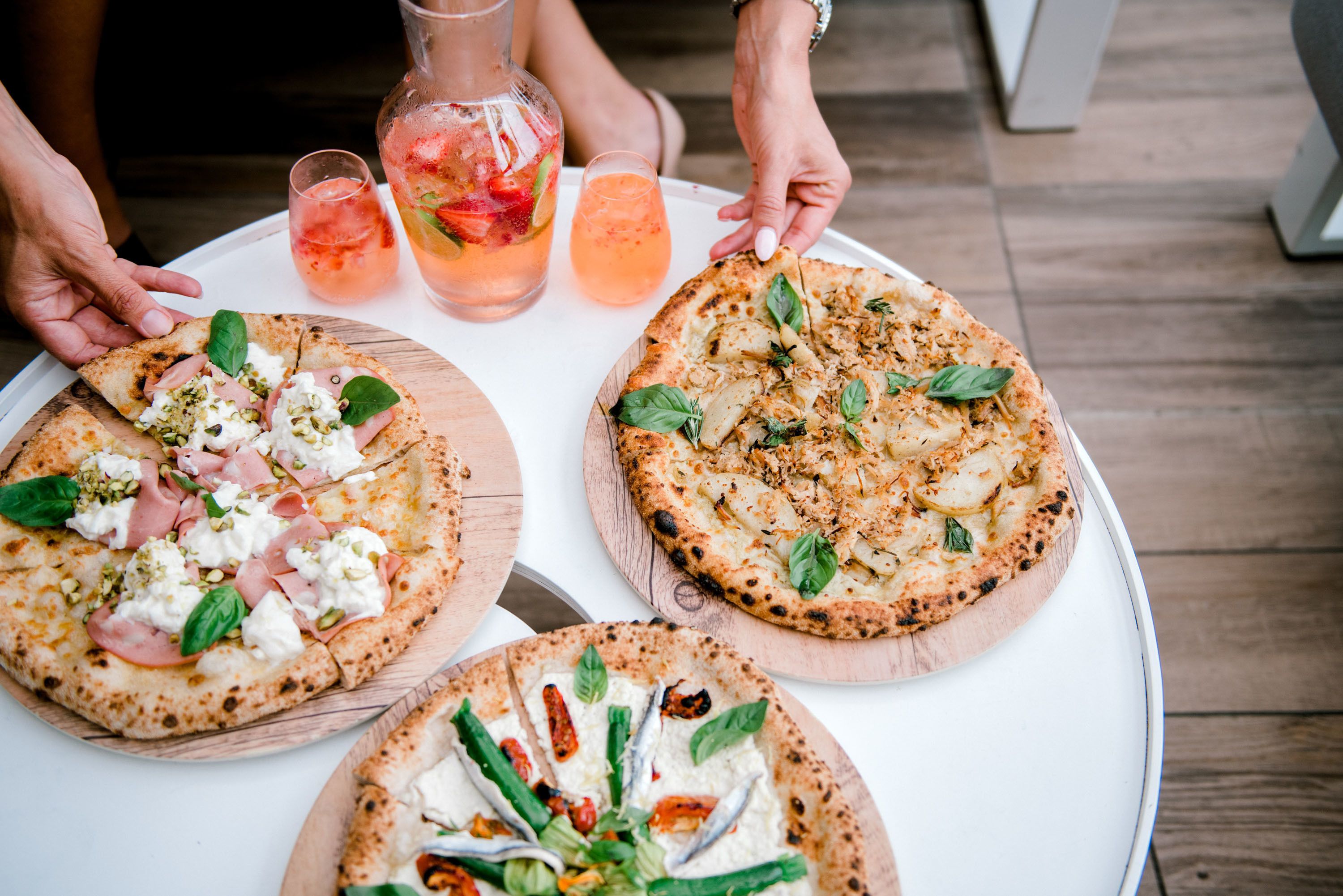 Drei Pizzen auf Holzbrettern mit unterschiedlichen Belägen, ein Eisteekrug und zwei Gläser eines rosa Getränks auf einem runden Tisch.
