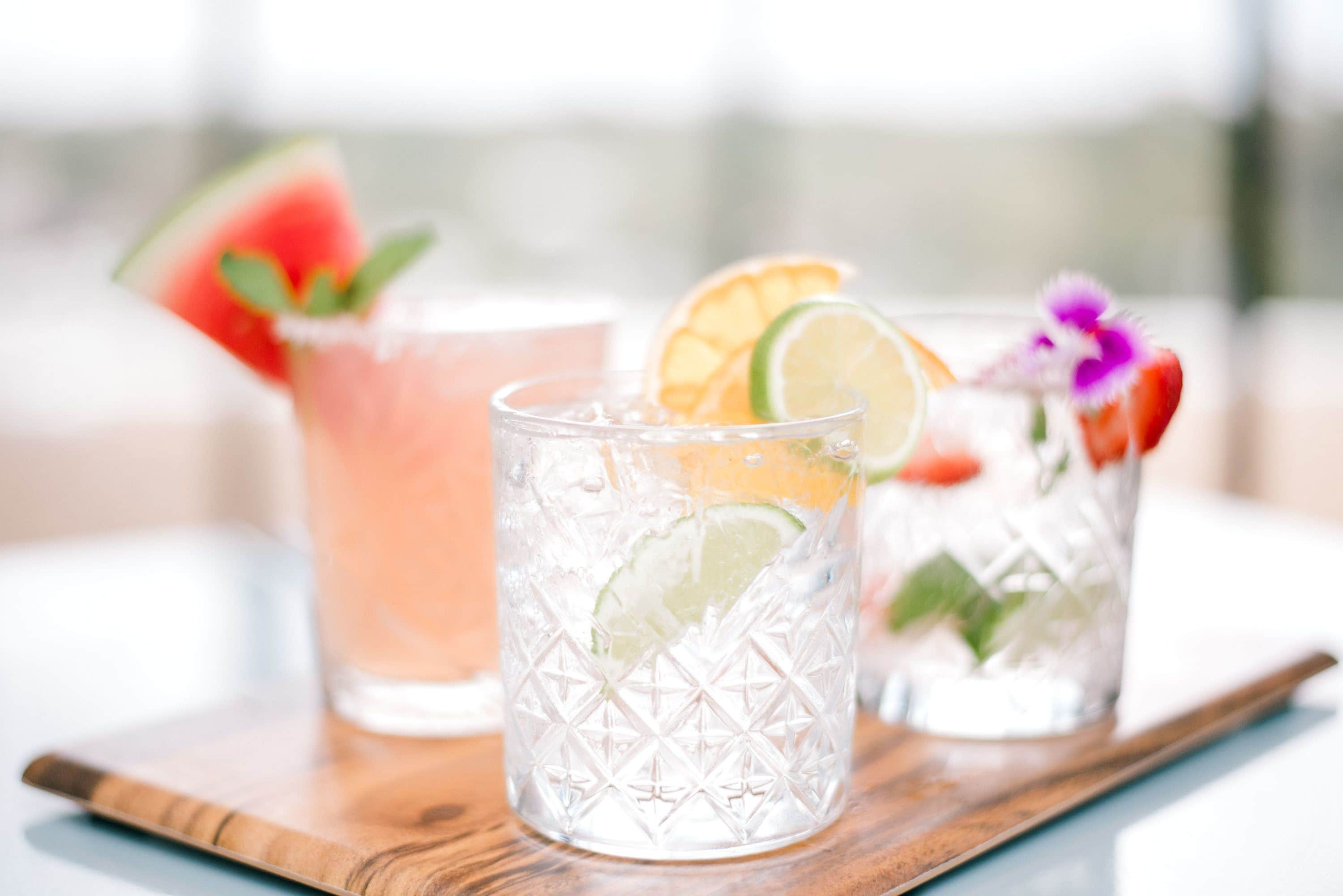 Tre glas med iskalla drycker, garnerade med lime och fruktskivor, på ett träbricka.