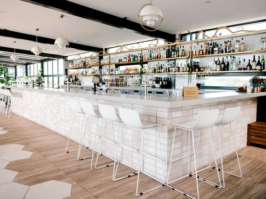 A bar area with a long counter, white bar stools, and shelves stocked with various bottles.