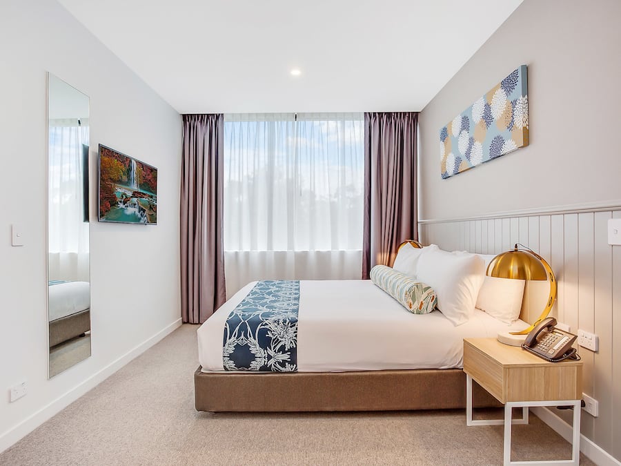 A modern hotel room with a large bed, a bedside table with a lamp, and a view of the city through large windows.