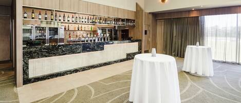 A hotel bar with a marble countertop, multiple shelves of liquor bottles, and two round tables with white tablecloths.