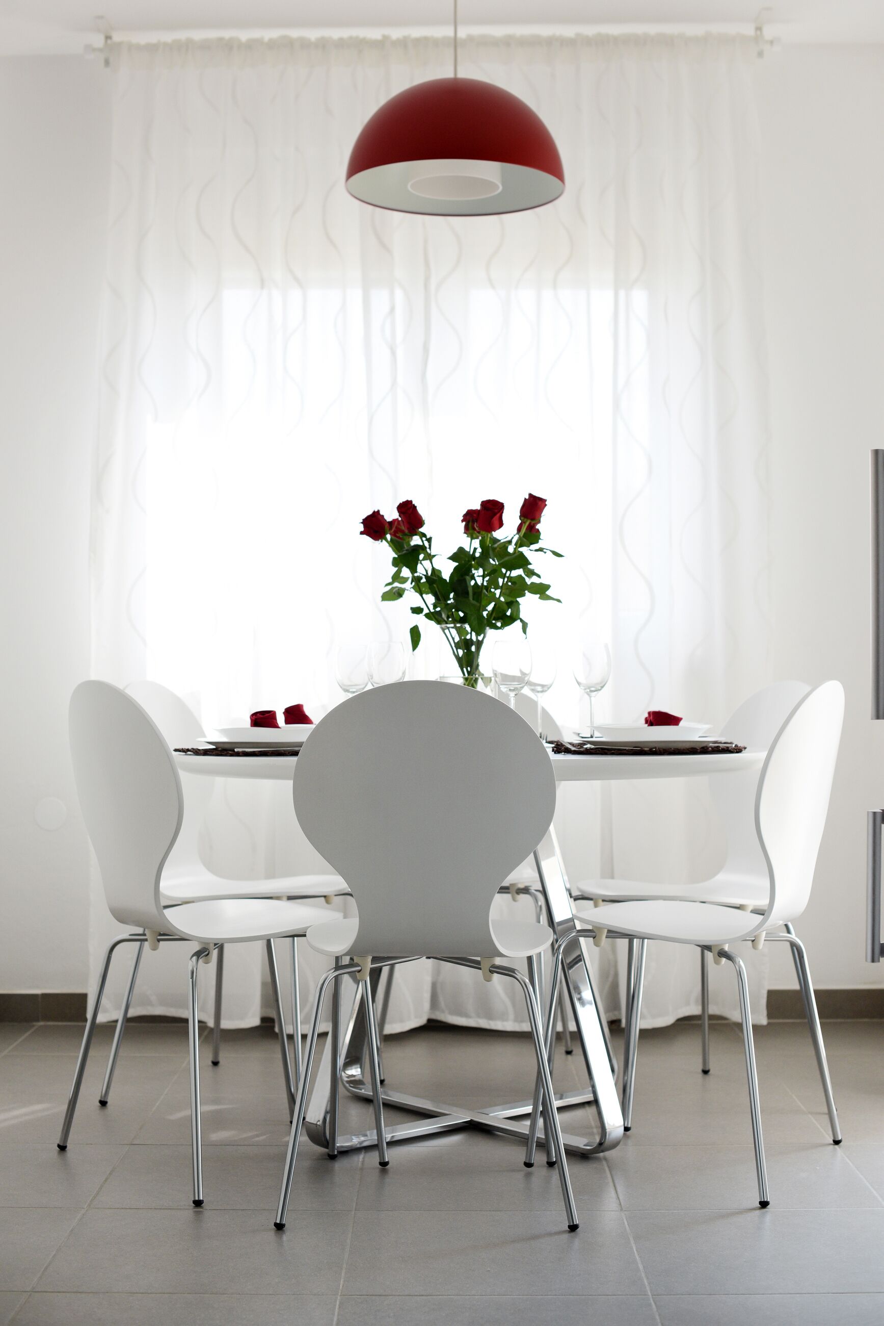 A dining area with a white table, four white chairs, a red pendant light, and a vase with red roses.