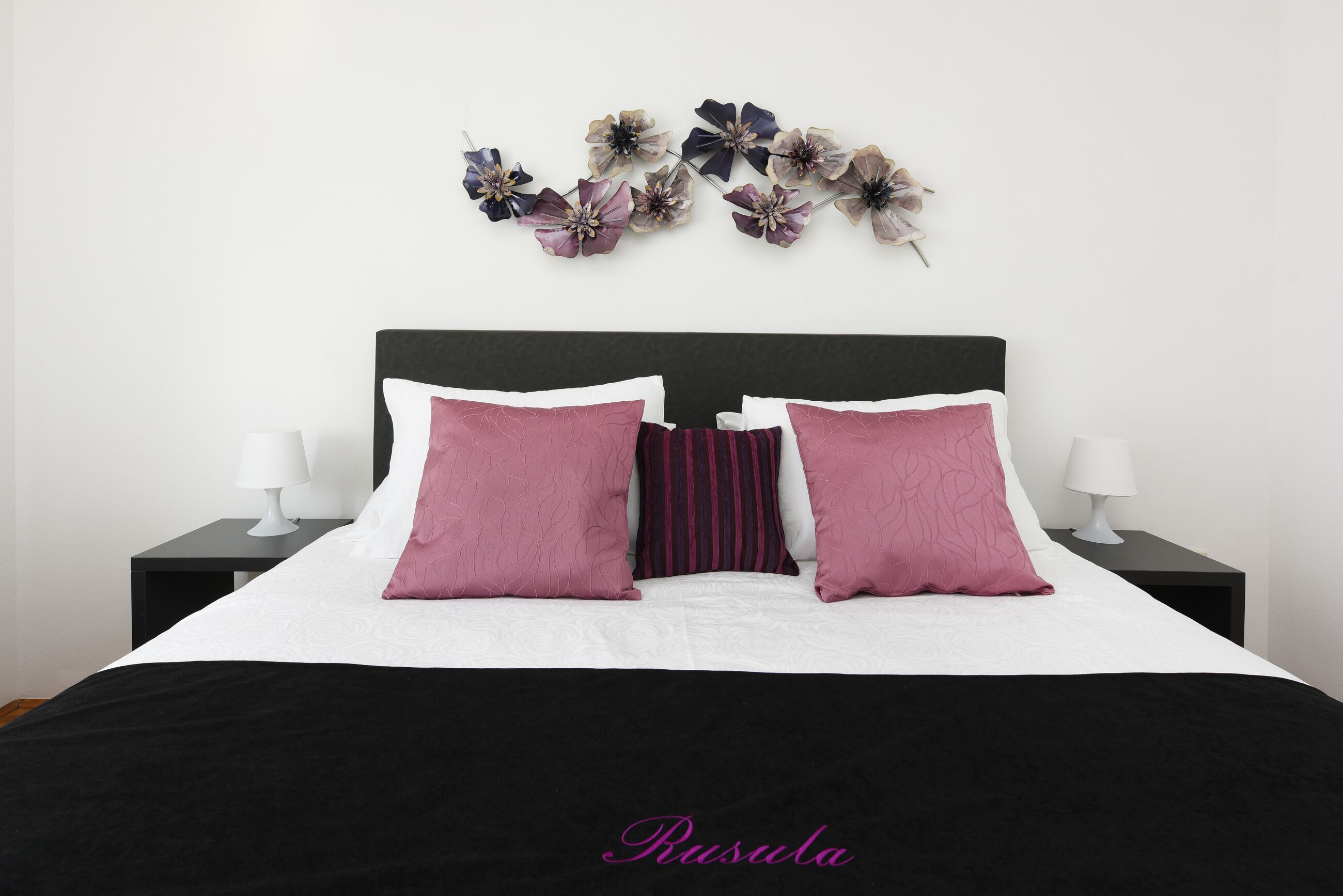 A neatly made bed with a black headboard, white and pink pillows, and a decorative floral arrangement above it.