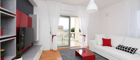 Een moderne woonkamer met een witte bank, rode accenten, een flatscreen televisie en een balkon met tuinmeubilair.