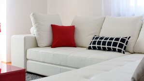 A white leather sofa with two pillows, one red and one black and white checkered, in a room with a red side table and a white curtain. - Apartment Rusula Red (Zadar)