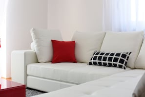 Un canapé blanc avec deux coussins, l’un rouge et l’autre à motifs noirs et blancs, dans une pièce où se trouvent une table rouge et un mur blanc.
