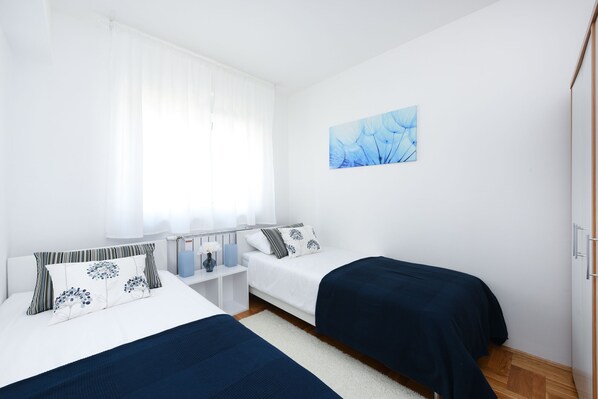 A bedroom with two beds, a white wardrobe, a bedside table with a lamp, and a window with sheer curtains.