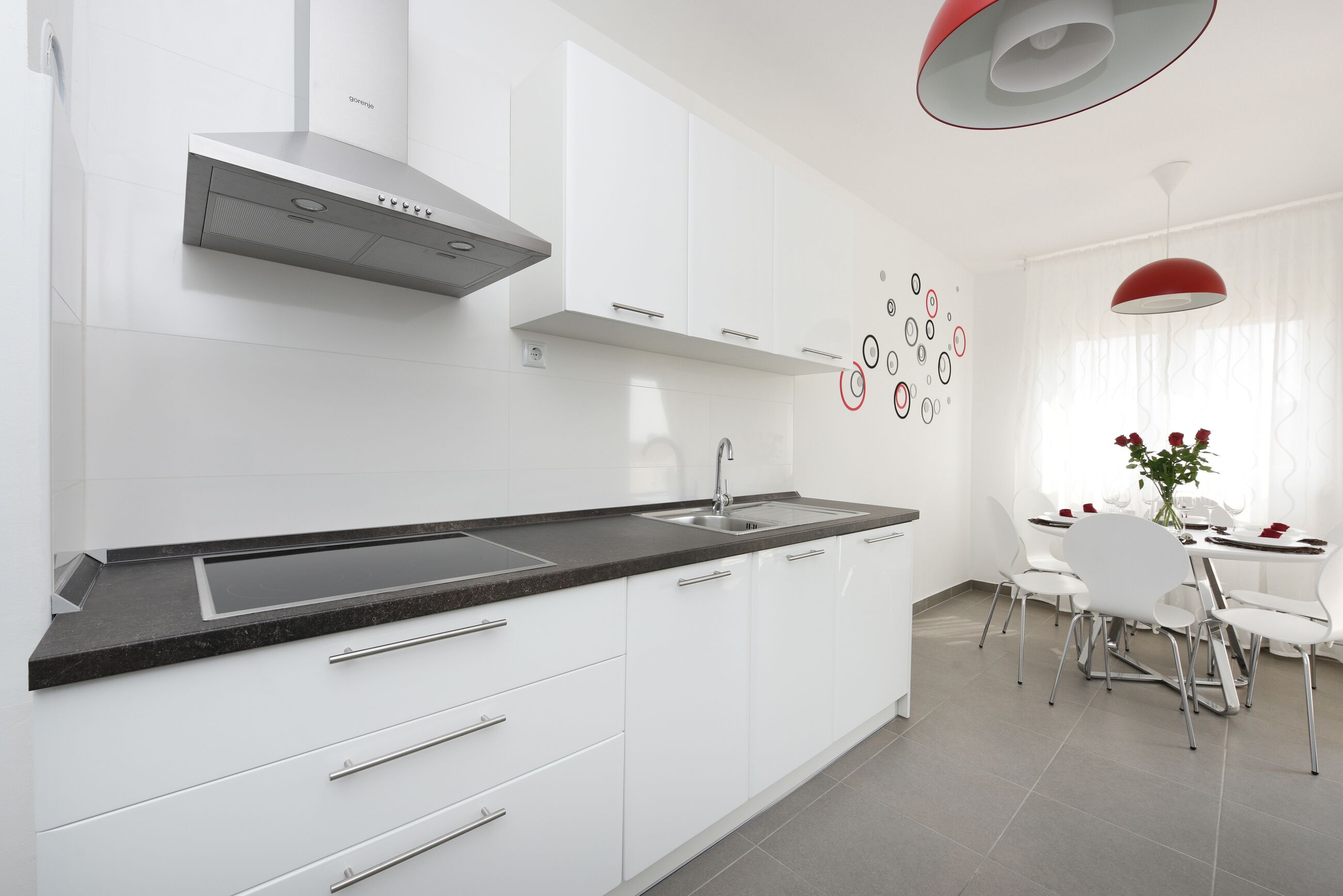 A modern kitchen with white cabinets, a black countertop, and a stainless steel range hood. There is a dining area with a white table and chairs, and a red pendant light above.